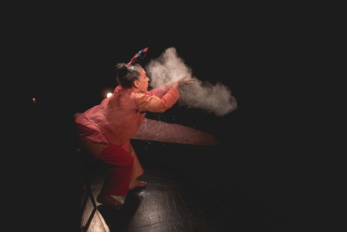 Imagen con fondo y piso negro, en el centro, está Carmen, bailarina de la obra de pelo canoso, lleva moño en forma de tomate y un remolino de papel.  Está semi agachada de perfil soplando de sus manos, un polvillo blanco que se suspende  en el aire.
