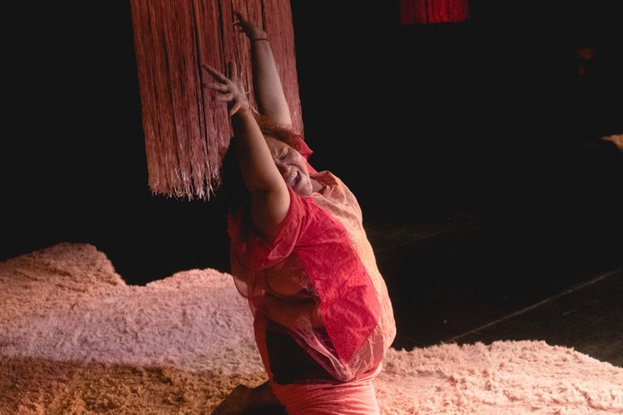 Imagen en fondo negro cuelga una lámpara con tiras finas en color rosado sobre una alfombra felpuda está marcela bailarina de la obra que aparece vestida de rosado y cuerpo voluminoso, está sobre sus rodillas con ambos brazos extendidos rostro inclinado hacia hombro derecho con expresión de goce y felicidad.